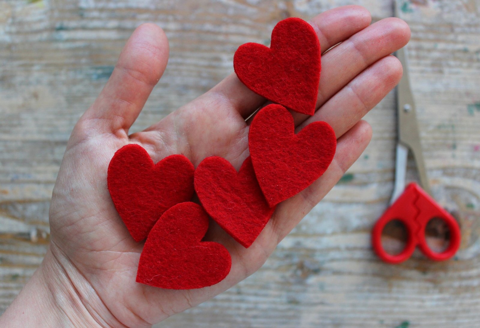 Hand holding five red felt hearts cut out with small red craft scissors on a wooden table